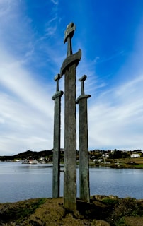 Three large bronze sword sculptures stand upright on a rock by the water. The background includes a tranquil body of water and a distant view of a small town with hills and houses. The sky is partly cloudy with blue and white hues.