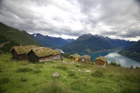 A scenic view of rustic cabins with grassy roofs nestled in a lush mountainous landscape. The foreground features a green meadow with scattered rocks, while the background showcases a tranquil fjord surrounded by towering mountains under a cloudy sky.
