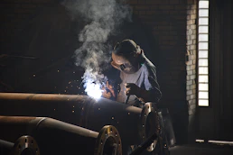 Industrial scene showing workers welding large steel pipes with sparks flying in a dark, moody setting