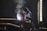 A worker wearing protective gear and a welding mask is engaged in welding large metal pipes in an industrial setting. Bright sparks and smoke are emanating from the point of contact, indicating active welding. The environment is dimly lit, with ambient light coming from a nearby window.