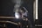 A worker wearing protective gear and a welding mask is engaged in welding large metal pipes in an industrial setting. Bright sparks and smoke are emanating from the point of contact, indicating active welding. The environment is dimly lit, with ambient light coming from a nearby window.