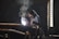 A worker wearing protective gear and a welding mask is engaged in welding large metal pipes in an industrial setting. Bright sparks and smoke are emanating from the point of contact, indicating active welding. The environment is dimly lit, with ambient light coming from a nearby window.