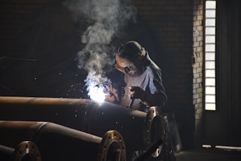 A worker wearing protective gear and a welding mask is engaged in welding large metal pipes in an industrial setting. Bright sparks and smoke are emanating from the point of contact, indicating active welding. The environment is dimly lit, with ambient light coming from a nearby window.