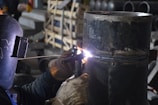 A welder wearing protective gloves and a helmet is working on a large metal pipe. Sparks and bright light are visible where the welding is taking place. The background contains various industrial materials and equipment.
