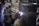 A welder wearing protective gloves and a helmet is working on a large metal pipe. Sparks and bright light are visible where the welding is taking place. The background contains various industrial materials and equipment.
