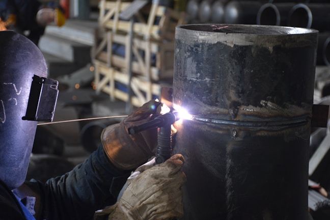 A welder wearing protective gloves and a helmet is working on a large metal pipe. Sparks and bright light are visible where the welding is taking place. The background contains various industrial materials and equipment.