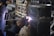 A welder wearing protective gloves and a helmet is working on a large metal pipe. Sparks and bright light are visible where the welding is taking place. The background contains various industrial materials and equipment.