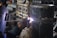 A welder wearing protective gloves and a helmet is working on a large metal pipe. Sparks and bright light are visible where the welding is taking place. The background contains various industrial materials and equipment.