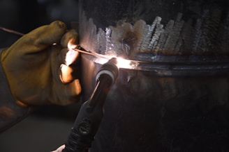 A close-up view of a welding process, showing a gloved hand holding a welding torch against a metal surface. Bright sparks and a small, intense light indicate active welding. The metal surface displays molten areas and welding marks, highlighted by the light from the torch.