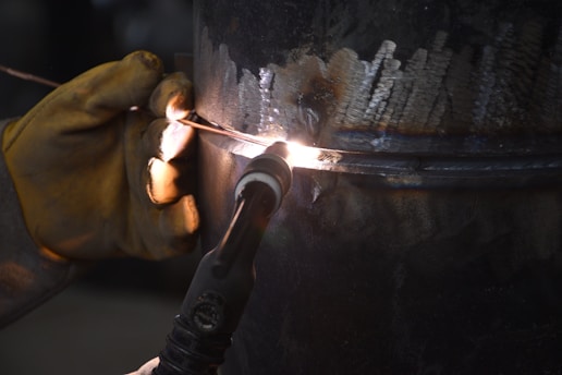 A close-up view of a welding process, showing a gloved hand holding a welding torch against a metal surface. Bright sparks and a small, intense light indicate active welding. The metal surface displays molten areas and welding marks, highlighted by the light from the torch.