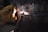 A close-up view of a welding process, showing a gloved hand holding a welding torch against a metal surface. Bright sparks and a small, intense light indicate active welding. The metal surface displays molten areas and welding marks, highlighted by the light from the torch.