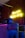 A wooden wall illuminated by a neon yellow sign that reads 'Camora Barber Shop'. Below the sign are metal barrels and lockers adorned with colorful posters. A glittery motorcycle helmet is positioned on top of one of the lockers, next to a sleek black helmet.