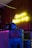 A wooden wall illuminated by a neon yellow sign that reads 'Camora Barber Shop'. Below the sign are metal barrels and lockers adorned with colorful posters. A glittery motorcycle helmet is positioned on top of one of the lockers, next to a sleek black helmet.