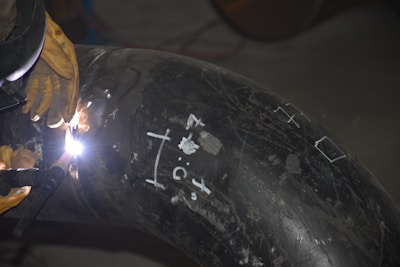 A welder is working on a large metal pipe, using a welding torch. The pipe is marked with white chalk symbols and numbers. The welder is wearing protective gloves and the bright welding light illuminates the immediate area.