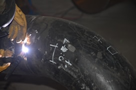 A welder is working on a large metal pipe, using a welding torch. The pipe is marked with white chalk symbols and numbers. The welder is wearing protective gloves and the bright welding light illuminates the immediate area.