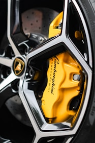 A close-up view of a car wheel showcasing a sleek black and silver alloy rim design, with a prominent yellow brake caliper featuring the logo of a renowned automotive brand. The disc rotor is visible behind the caliper.