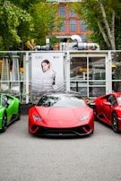 A red sports car is prominently displayed in the center, flanked by two other sports cars, one green and one red. The background features a large poster of a woman and a building with glass windows and brickwork, partially obscured by trees.