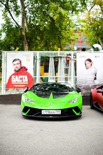 A bright green sports car is parked in front of a backdrop of trees and promotional posters. The car is predominantly green with black accents and has a visible license plate. To its side is another sports car partially visible in red. The background features posters of people, suggesting an event or advertisement setting.