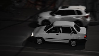 Two cars are driving on a road at night. The car in the foreground appears to be an older model, while a more modern SUV passes by in the background.