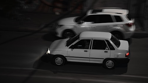 Two cars are driving on a road at night. The car in the foreground appears to be an older model, while a more modern SUV passes by in the background.