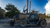 A large construction vehicle is parked on a worksite. It is positioned on a dirt area surrounded by trees and buildings. The sun is shining brightly in the background, creating a glare on a nearby building. The vehicle has yellow and black coloring and appears to be used for heavy lifting or digging.