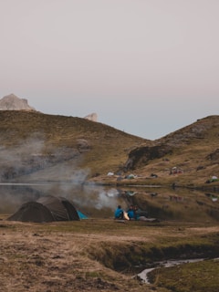 A serene campsite near a calm water body with several tents scattered across a grassy landscape. Three people are sitting around a campfire in the foreground, surrounded by a few more tents and distant hills. The gentle smoke from the campfire rises into the evening sky, creating a peaceful, outdoor setting.