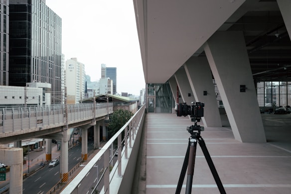 A modern urban setting with a tripod-mounted camera on a spacious, concrete balcony or walkway. The scene overlooks a busy road with several vehicles and a monorail track. Tall buildings in varying designs can be seen in the background, suggesting a cityscape.