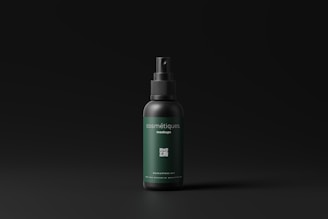 A cosmetic spray bottle with a dark green label and black cap is centered in a minimalistic setting. The label includes text and a small QR code. The background is a simple matte black, which highlights the product.