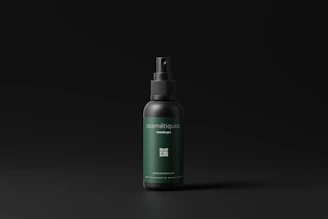 A cosmetic spray bottle with a dark green label and black cap is centered in a minimalistic setting. The label includes text and a small QR code. The background is a simple matte black, which highlights the product.
