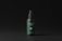 A cosmetic spray bottle with a dark green label and black cap is centered in a minimalistic setting. The label includes text and a small QR code. The background is a simple matte black, which highlights the product.