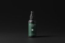 A cosmetic spray bottle with a dark green label and black cap is centered in a minimalistic setting. The label includes text and a small QR code. The background is a simple matte black, which highlights the product.