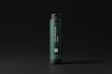 A dark green cylindrical bottle with a black cap stands against a dark background. The label on the bottle reads 'cosmétiques mockups' and includes a QR code as well as a website URL. The design is minimalistic and modern.