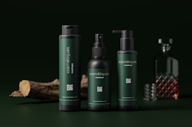 Three dark green cosmetic bottles with black caps are arranged in the foreground. Each bottle has the word 'cosmétiques' written on it. In the background, there is a glass decanter with a patterned design, partially filled with a brown liquid. A piece of wood is lying on the left side, adding a natural element.