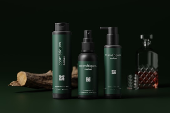 Three dark green cosmetic bottles with black caps are arranged in the foreground. Each bottle has the word 'cosmétiques' written on it. In the background, there is a glass decanter with a patterned design, partially filled with a brown liquid. A piece of wood is lying on the left side, adding a natural element.