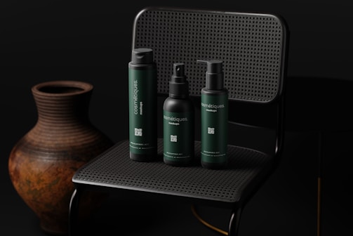 Three dark-colored cosmetic bottles are arranged on a dark, woven chair with an earthy-toned pottery vase next to it. The lighting is subdued, creating a sophisticated and sleek atmosphere.