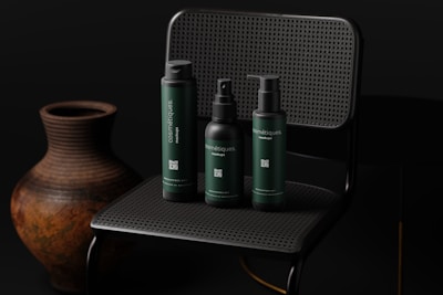 Three dark-colored cosmetic bottles are arranged on a dark, woven chair with an earthy-toned pottery vase next to it. The lighting is subdued, creating a sophisticated and sleek atmosphere.