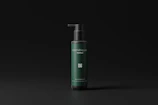 A sleek, dark green bottle with a black pump dispenser is set against a matte black background. The bottle features minimalist text branding, and the overall design exudes elegance and simplicity.