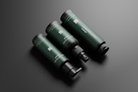 Three dark green cylindrical bottles with black caps and dispensers are arranged diagonally against a dark gray background. The bottles have 'cosmétiques mockup' written on them in white text along with QR codes and other fine text details.