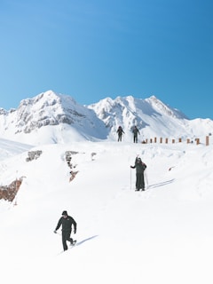 Several people are trekking across a snowy mountain landscape. The clear blue sky contrasts with the white snow-covered mountains in the background. The trekkers are wearing dark clothing and carrying poles, maneuvering through the snow.