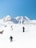 Several people are trekking across a snowy mountain landscape. The clear blue sky contrasts with the white snow-covered mountains in the background. The trekkers are wearing dark clothing and carrying poles, maneuvering through the snow.