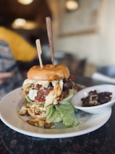 A lavish burger is placed on a white plate with a side dish of roasted mushrooms. The burger is piled high with various toppings including lettuce, tomatoes, cheese, and a patty, all enclosed in a bun pierced with wooden sticks. Melted cheese and sauce are dripping out of the burger. The background is out of focus but shows a person in a yellow shirt and blurred indoor setting.