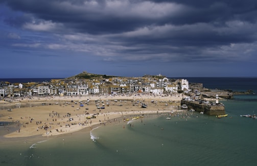 A coastal town with a sandy beach filled with people enjoying various activities. The town's buildings are closely packed, featuring a mix of traditional and modern styles. A small harbor with boats is visible, and the sky is overcast with dark clouds, suggesting an impending storm.