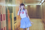 A person with long hair wearing a sailor-style outfit, featuring a blue skirt and white top with a red and blue collar. They are standing in a corridor with colorful tiled walls and appear to be holding a hand to their ear, suggesting they are listening intently.