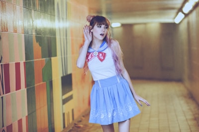 A person with long hair wearing a sailor-style outfit, featuring a blue skirt and white top with a red and blue collar. They are standing in a corridor with colorful tiled walls and appear to be holding a hand to their ear, suggesting they are listening intently.