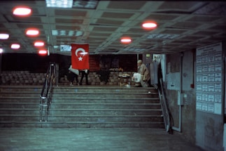 In an underground passage, a Turkish flag hangs prominently. Two people walk up the stairs to the left, while a person carrying a bag stands near the wall to the right. The ceiling features evenly spaced lights casting a red glow, and a board with notices is visible on the wall.