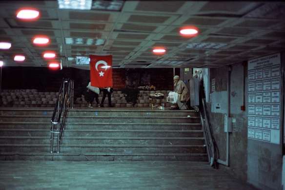 In an underground passage, a Turkish flag hangs prominently. Two people walk up the stairs to the left, while a person carrying a bag stands near the wall to the right. The ceiling features evenly spaced lights casting a red glow, and a board with notices is visible on the wall.