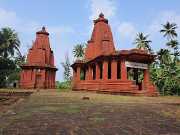 A traditional red sandstone temple structure with intricate carvings set amid lush greenery and coconut trees. The temple has two main towers, and a sign in a regional language above the entrance. The ground is covered in stone tiles with patches of grass.