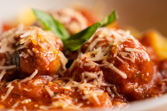 Meatballs covered in a rich tomato sauce topped with grated cheese are garnished with fresh basil. The dish appears appetizing and well-plated, with a focus on the textures of the cheese and sauce.