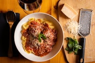 A bowl of pasta topped with a tomato-based sauce, garnished with grated cheese and basil leaves. Accompanying the dish is a glass of red wine, a block of cheese, a pile of grated cheese, basil leaves, and a cheese grater on a wooden cutting board.
