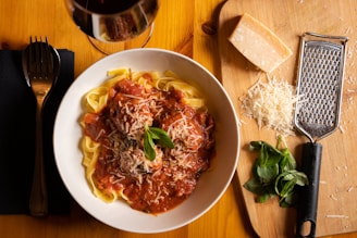 A bowl of pasta topped with a tomato-based sauce, garnished with grated cheese and basil leaves. Accompanying the dish is a glass of red wine, a block of cheese, a pile of grated cheese, basil leaves, and a cheese grater on a wooden cutting board.